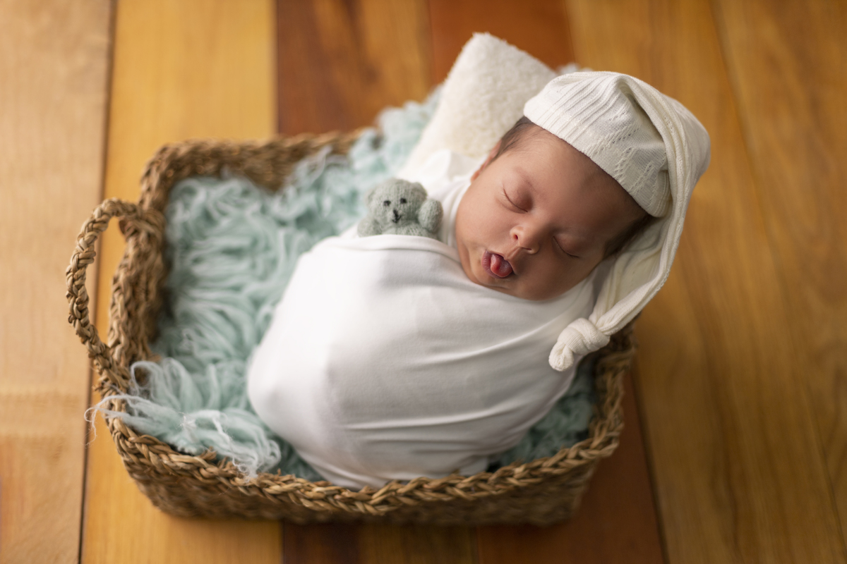 newborn coberto dentro na cesta com língua de fora e com gorro segurando ursinho de pelúcia
