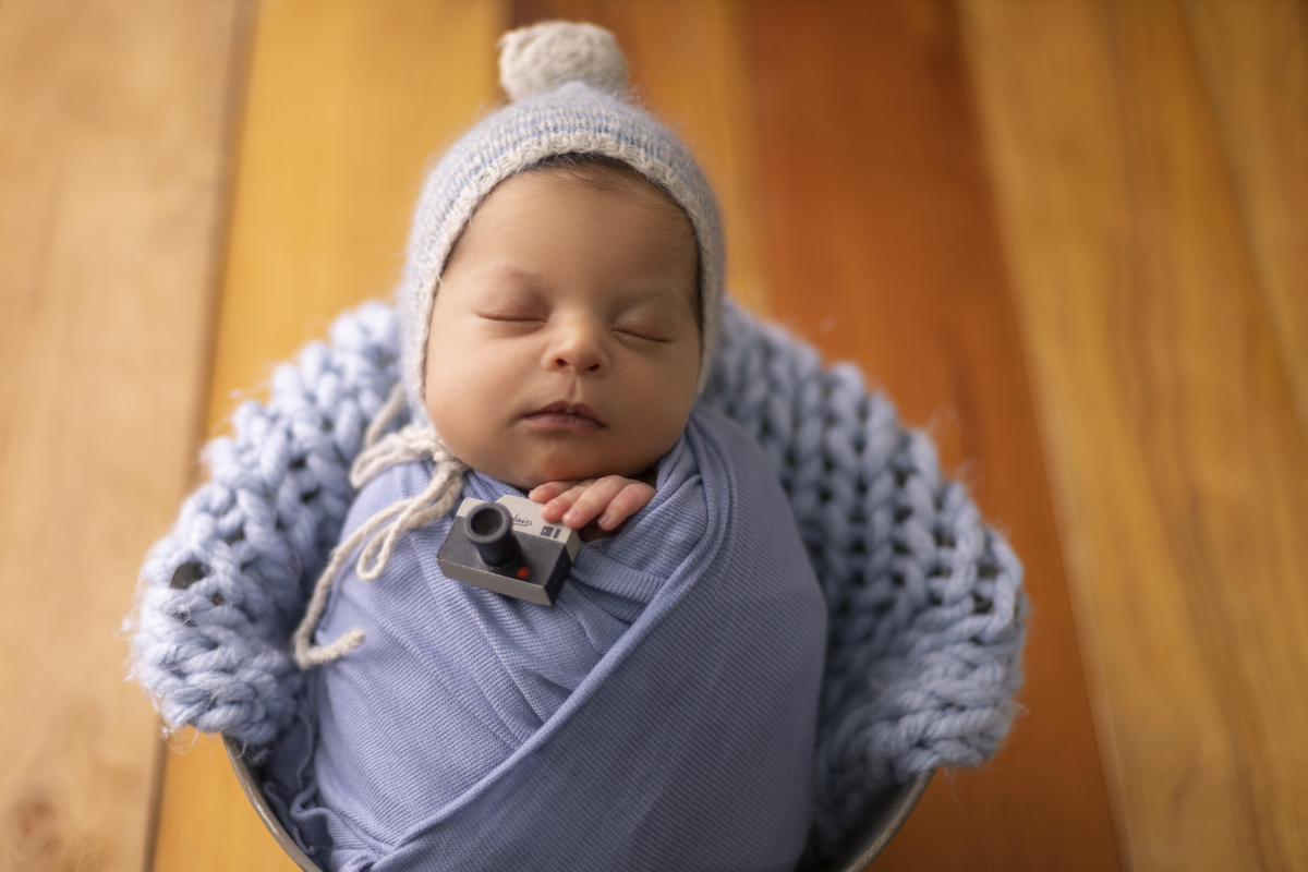 newborn coberto no balde marrom com gorro e segurando a câmera fotográfica