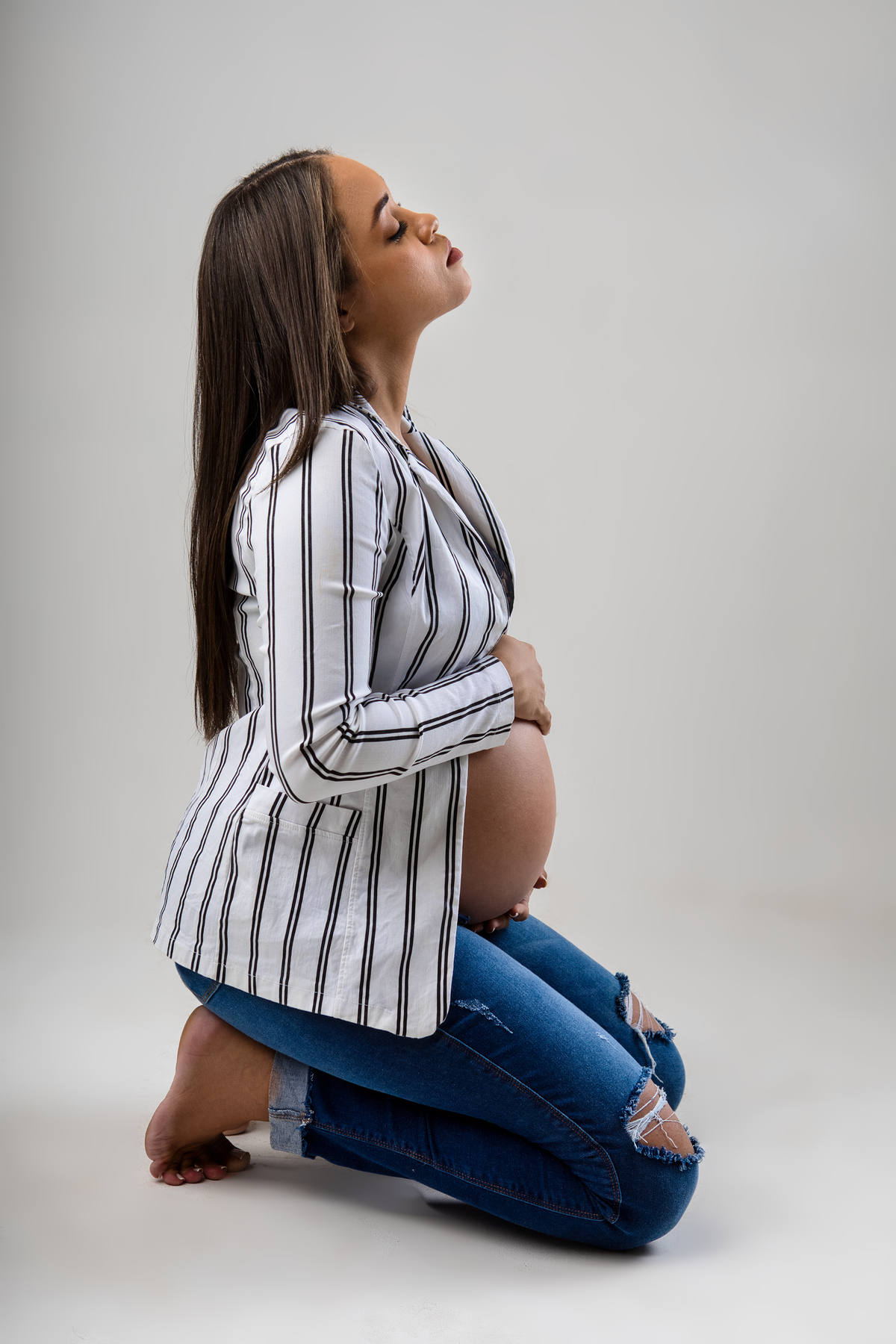 gestante de calça jeans e blazer ajoelhada abraçando a barriga 