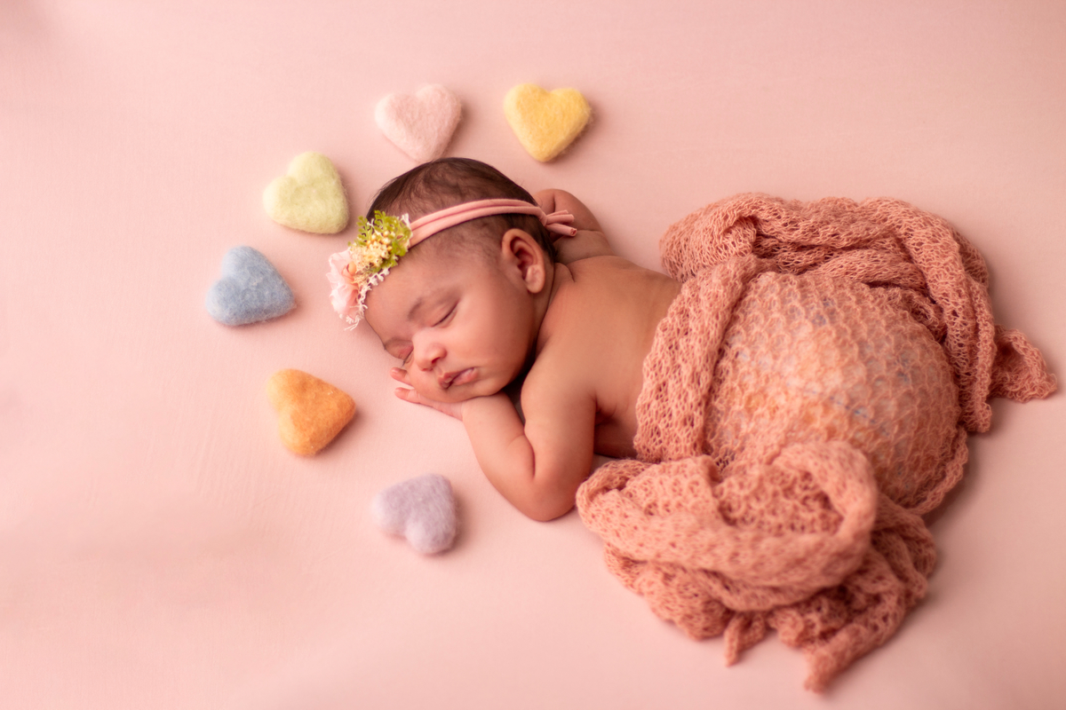 newborn de bruços com lençol bege, tiara e corações