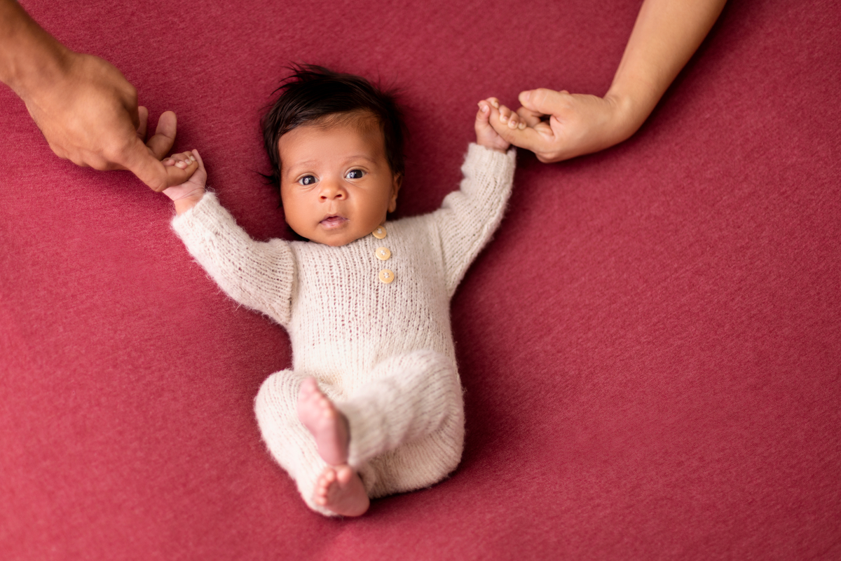 newborn de macaquinho deitado segurando as mãos de papai e mamãe
