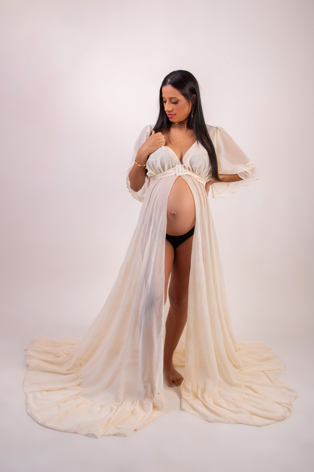 gestante com vestido longo branco com mão na cintura e outra segurando o vestido