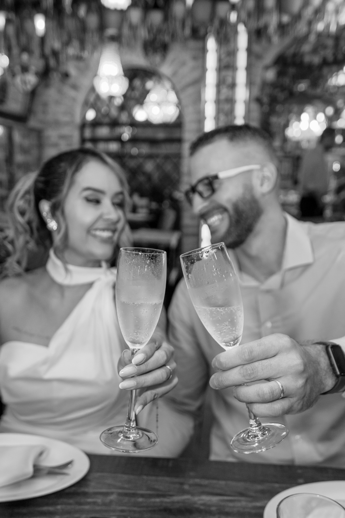 Foto muito tradicional, onde os noivos, o casal ficam desfocados no segundo plano com a expressão de extrema felicidade, e as taças no momento do brinde ficam em evidência.