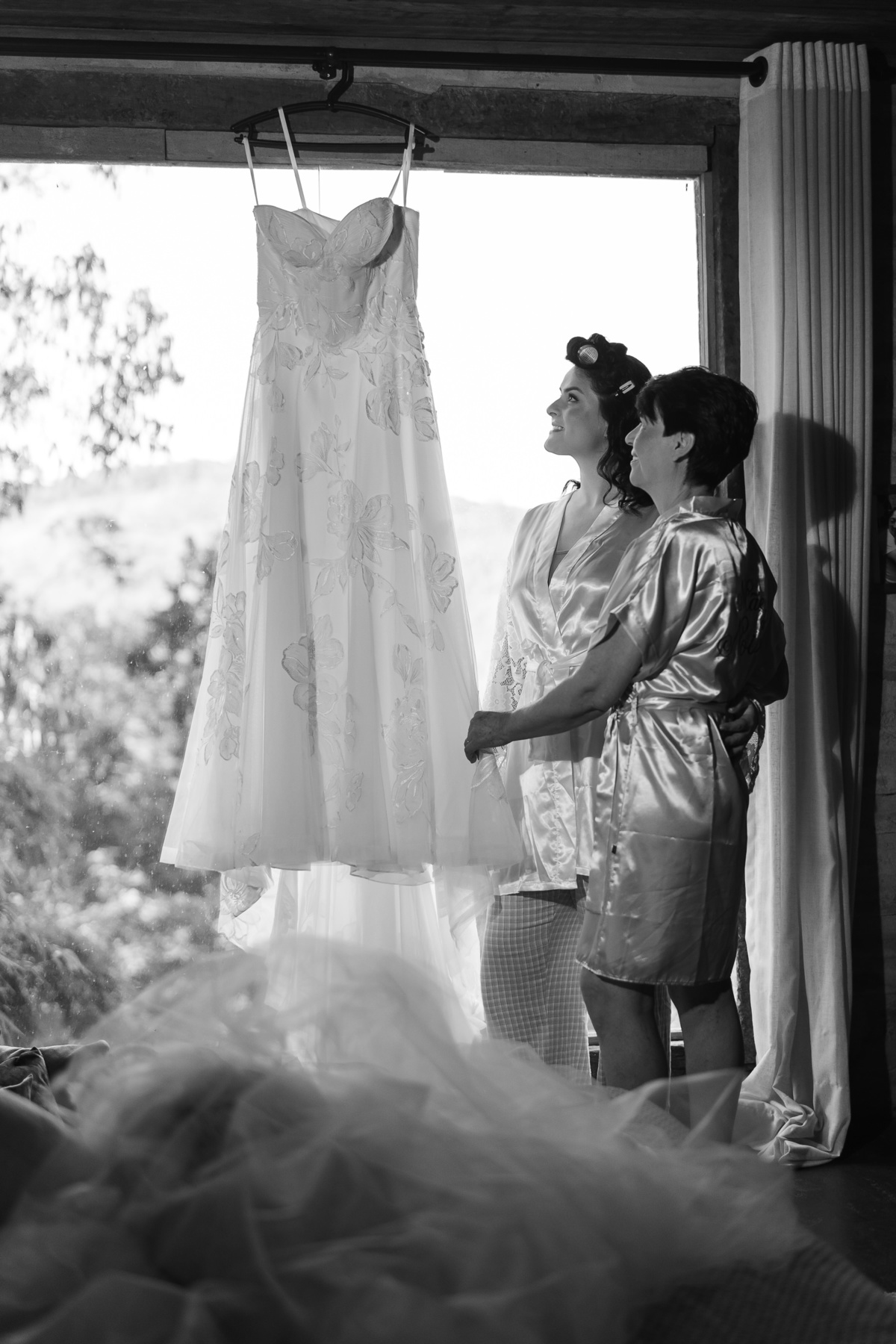 Foto da noiva e sua mãe admirando o seu vestido, pendurado em uma janela.