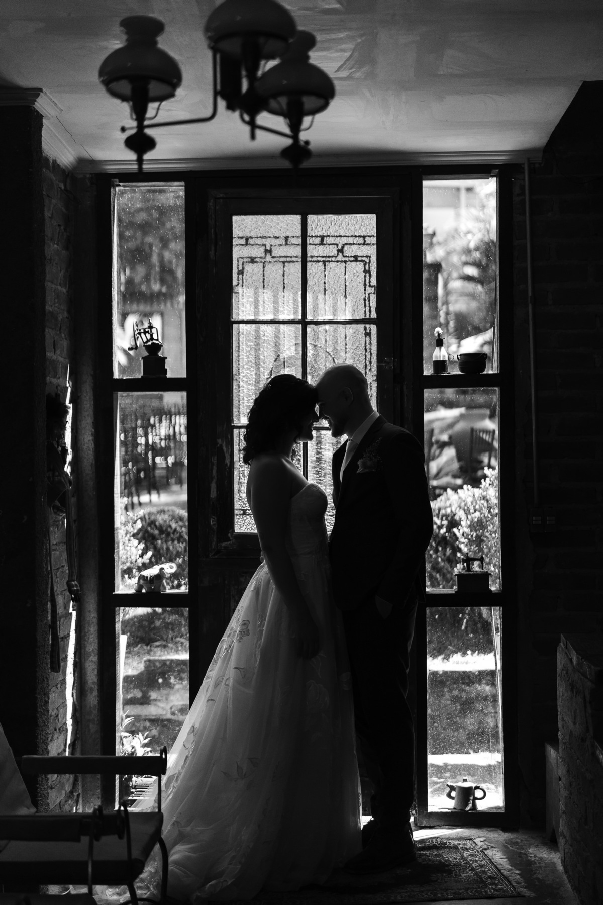 Foto artística, no estilo casamento no campo, que mostra somente a silhueta do casal, posicinado na contra luz em frente a uma porta de vidro.