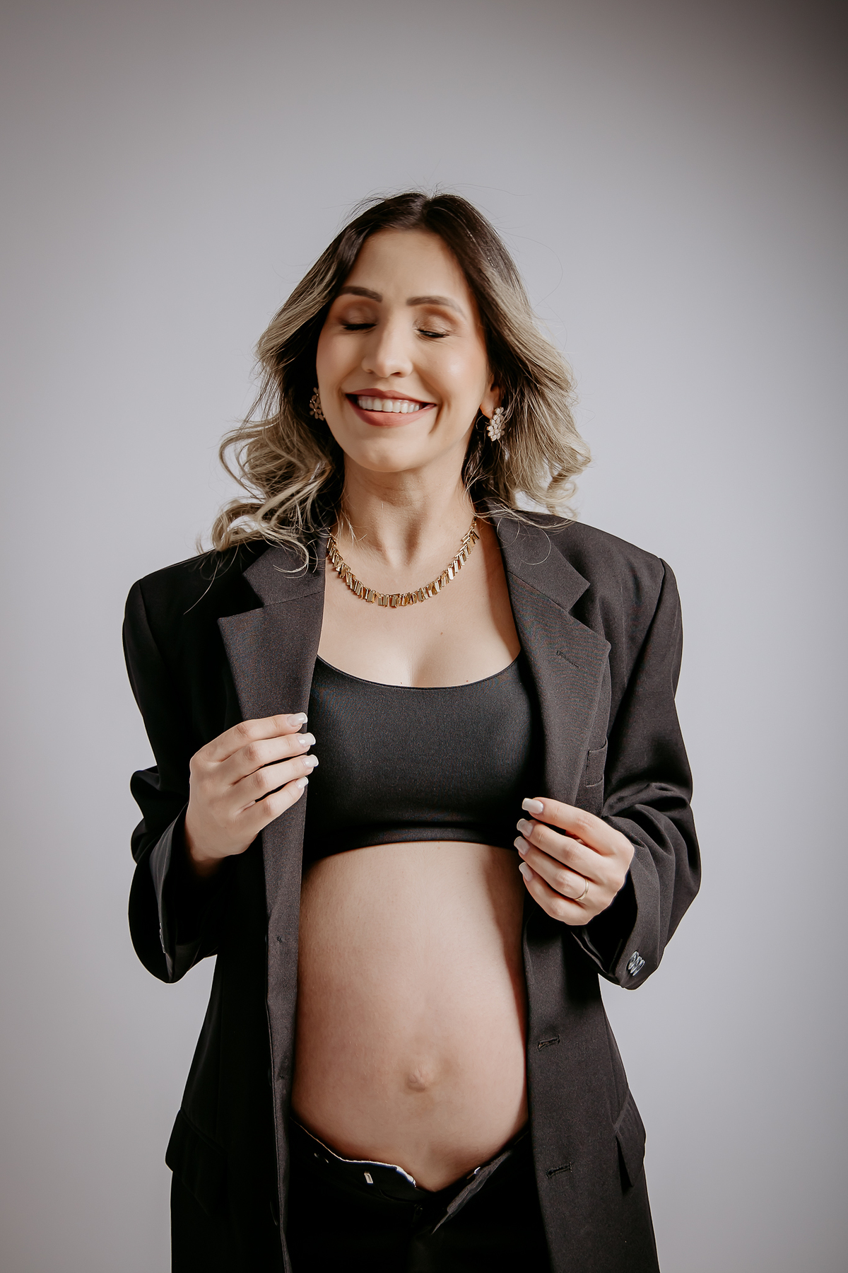 Gestante vestida de preto com blaser e calça preta e tope preto com as mãos delicadas no blaser com um sorriso muito feliz