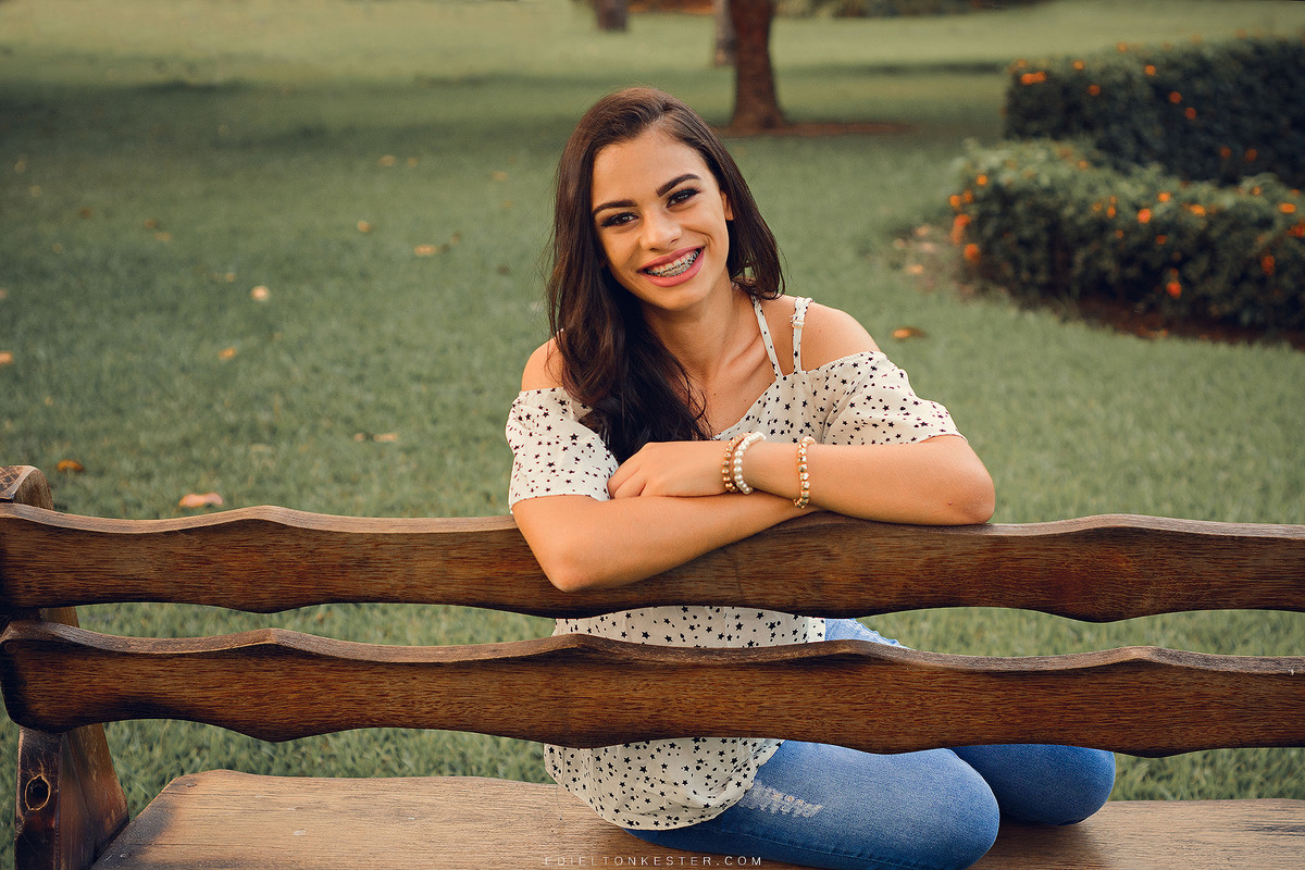 ensaio de 15 anos, menina sentada de costas no banco e sorrindo com braços cruzados