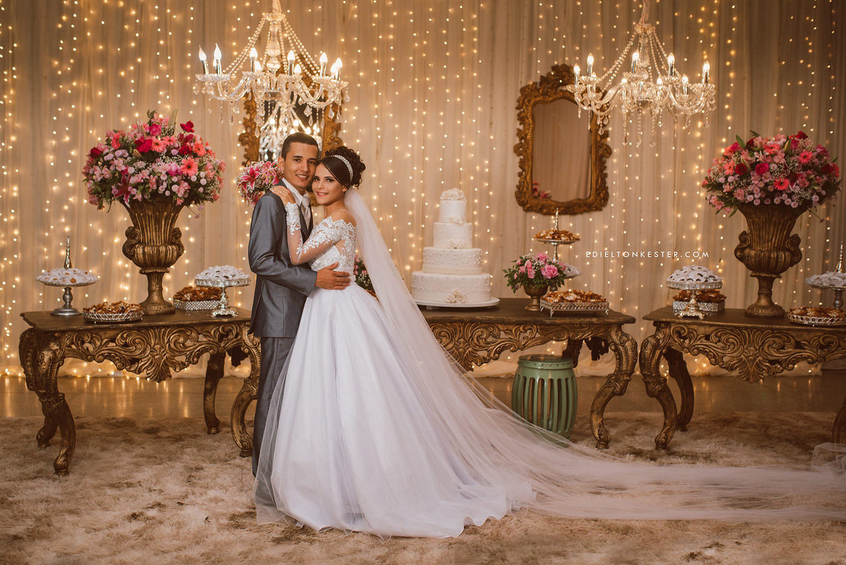 noivos posando para fotos em frente a mesa do bolo com muitas luzes véu da noiva grande e vestido lindo de noiva em ji paraná
