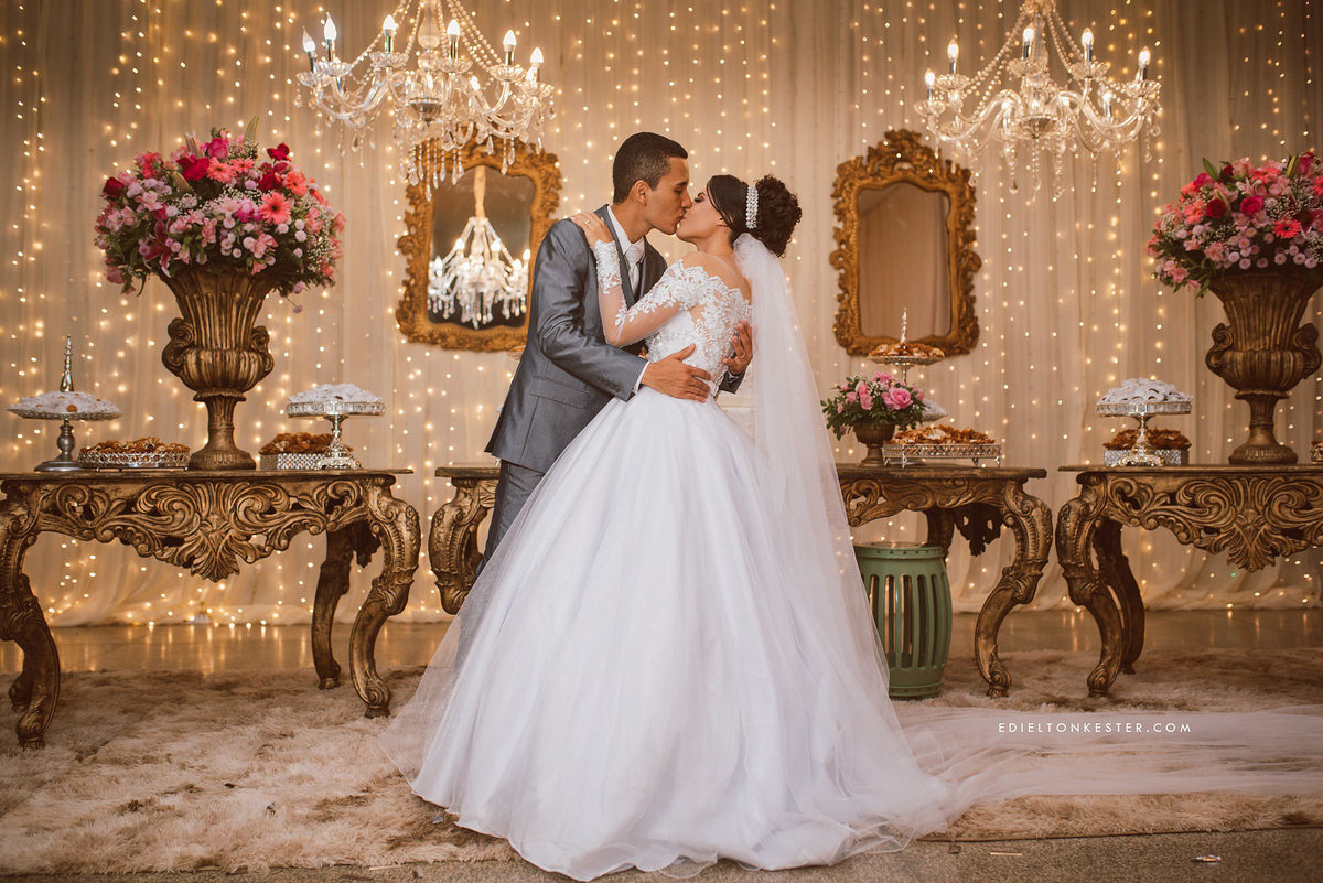 noivos posando para fotos em frente a mesa do bolo com muitas luzes véu da noiva grande e vestido lindo de noiva em ji paraná
