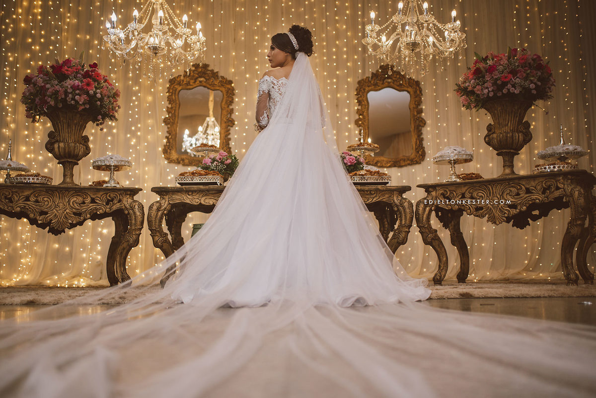 noiva de costas mostrando seu lindo vestido e véu grande em frente a mesa de doces em seu casamento