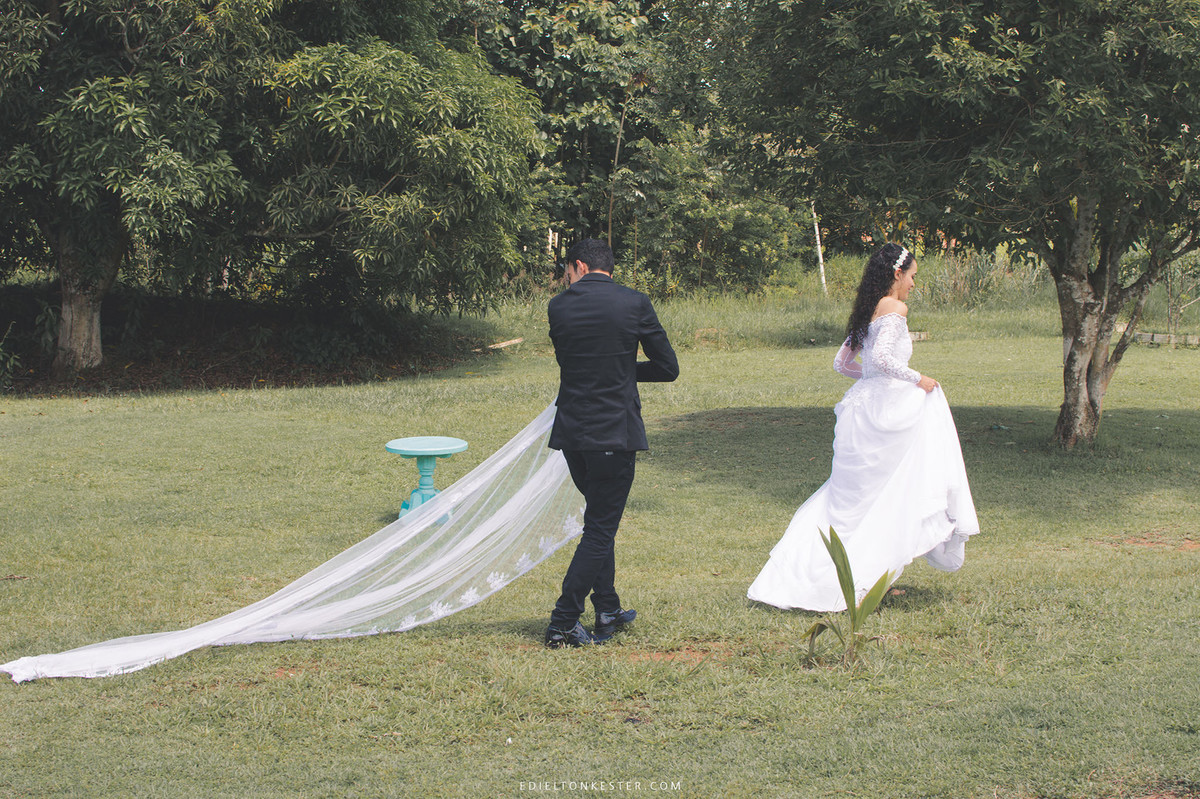 ensaio de casal, noivo segurando veu indo atras da noiva que caminham no gramado