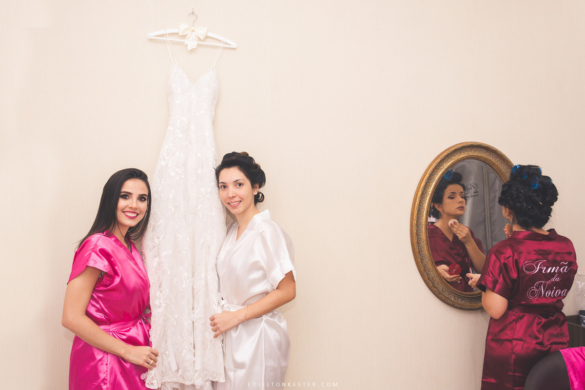 noiva e melhor amiga da noiva segurando vestido no making of enquanto a irma da noiva se maquia