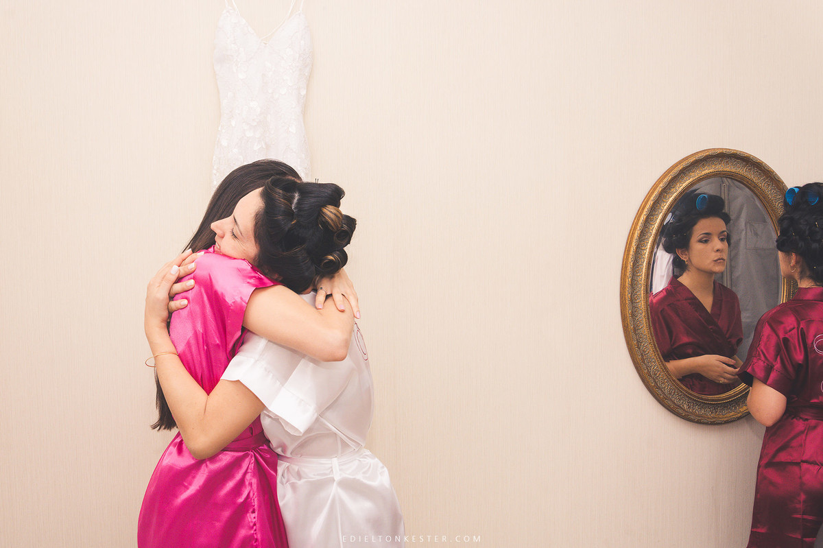 noiva e sua melhor amiga se abraçando e irma da noiva se maquia em frente ao espelho