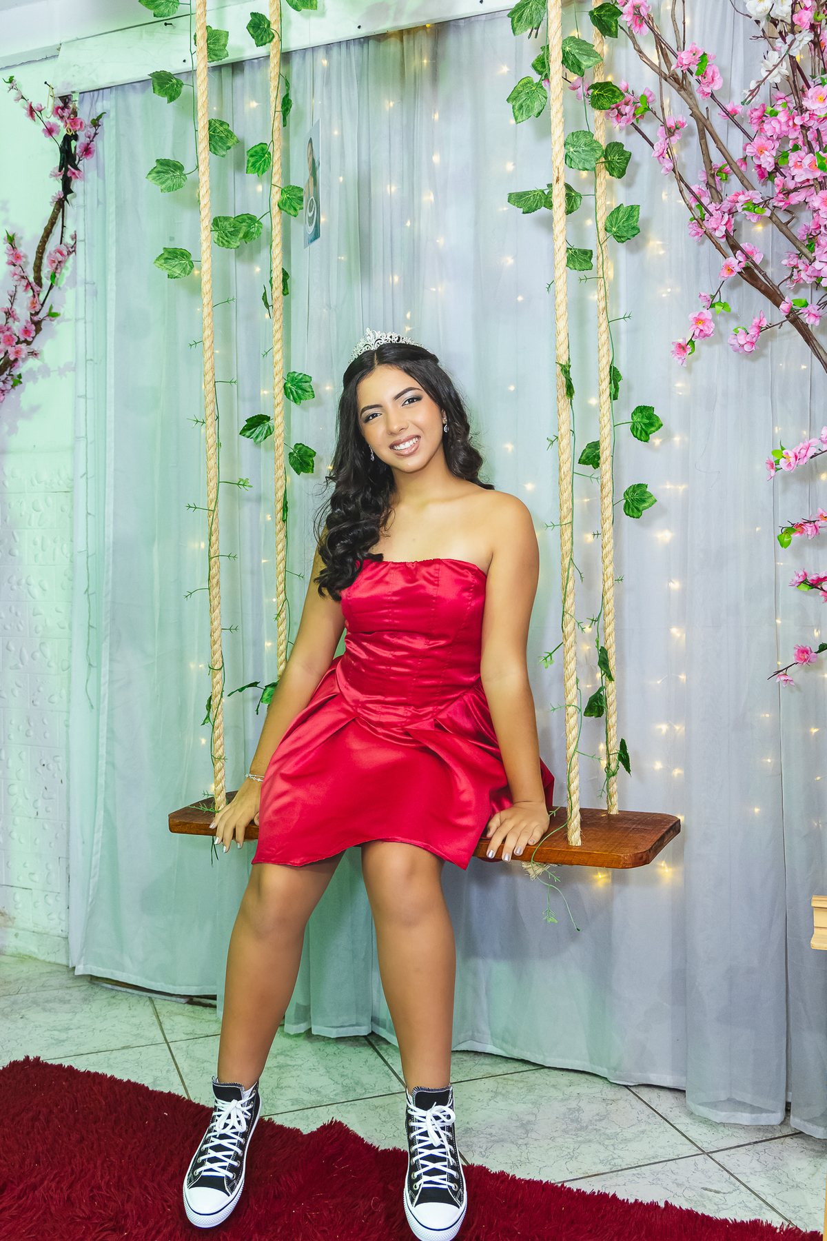 amin posa sorridente no balanço cenográfico, usando um elegante vestido vermelho e tiara, em um cenário delicado que remete a um verdadeiro conto de fadas.