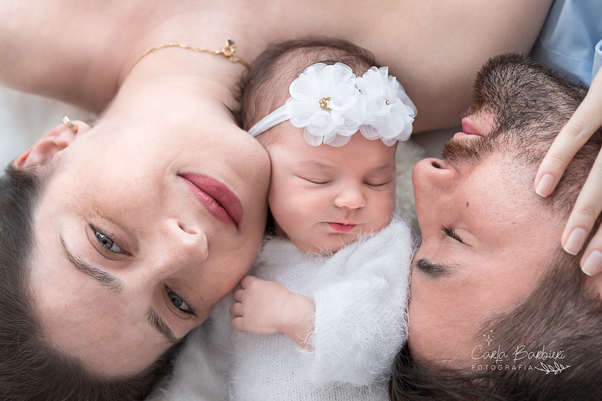 Fotografia da bebê Júlia com a mamãe Flávia  e o papai Marcelo deitados em tapete de pele branca.
