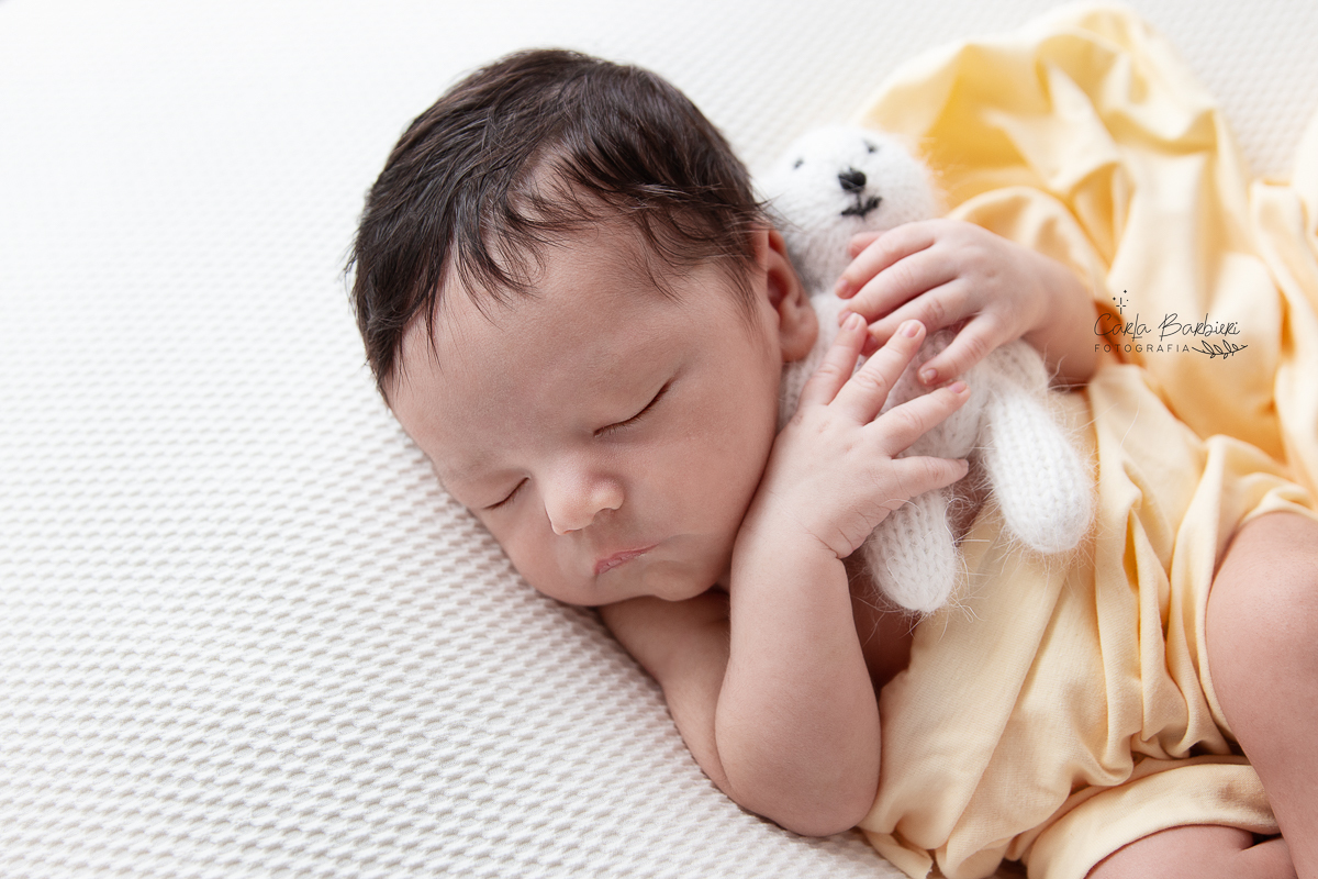 Close lateral do bebê Samuel em fotografia de barriga para cima coberto com wrap amarelo segurando um ursinho