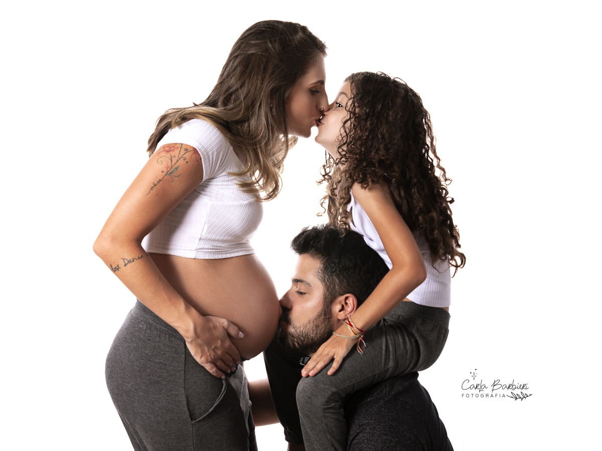 papai beijando a barriga da mamãe e a filha menor nos ombros do pai beijando a mamãe