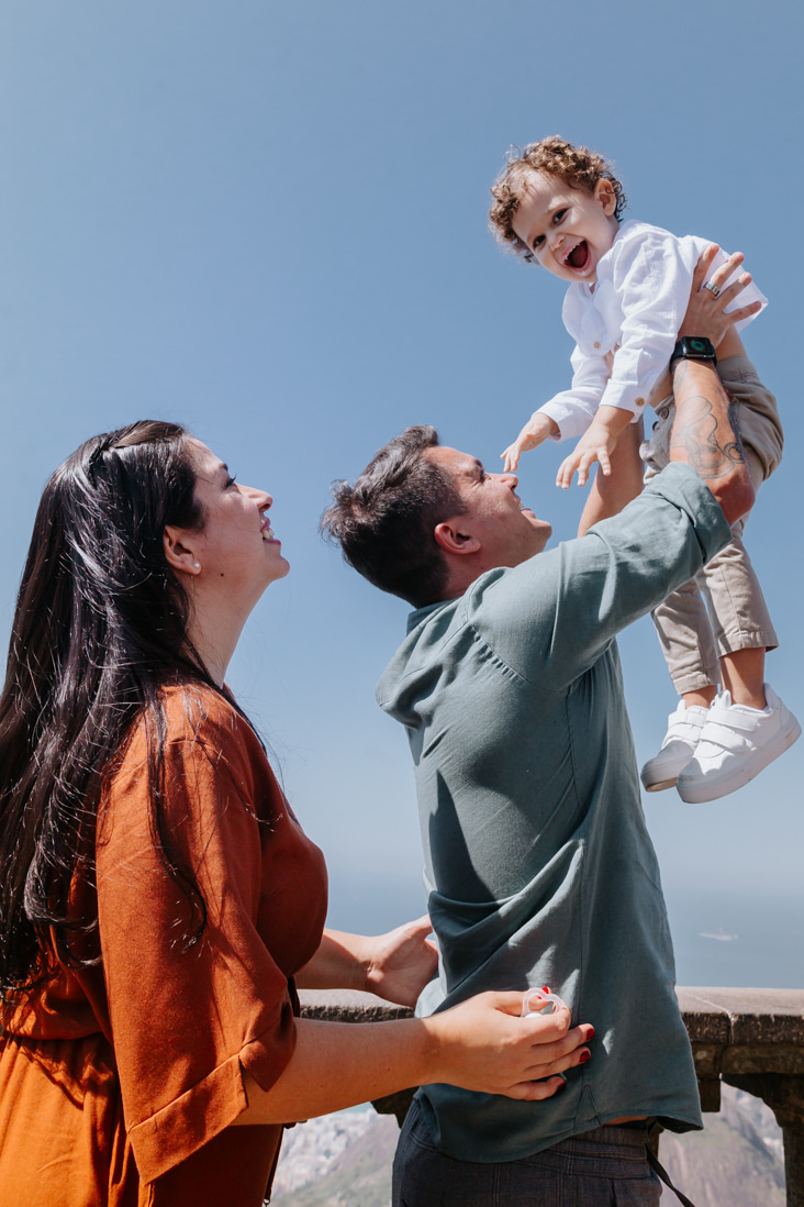 pai levanta criança apos batismo e mae atras dele observa e sorri