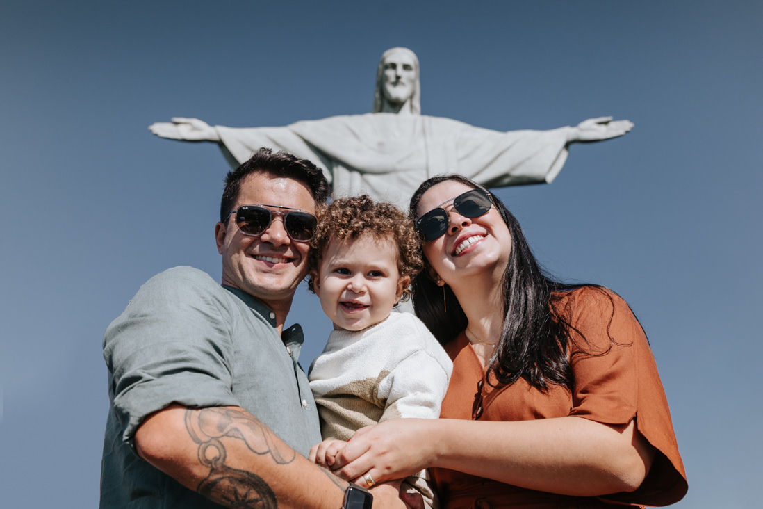 pai e mae de oculos escuros e  filho olhando para a camera no dia do batizado no cristo redentor
