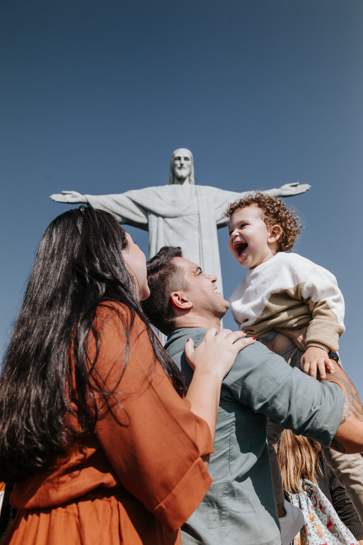 mae pai olham para o filho que é jogado para o alto com o com o cristo redentor ao fundo no dia do seu batizado