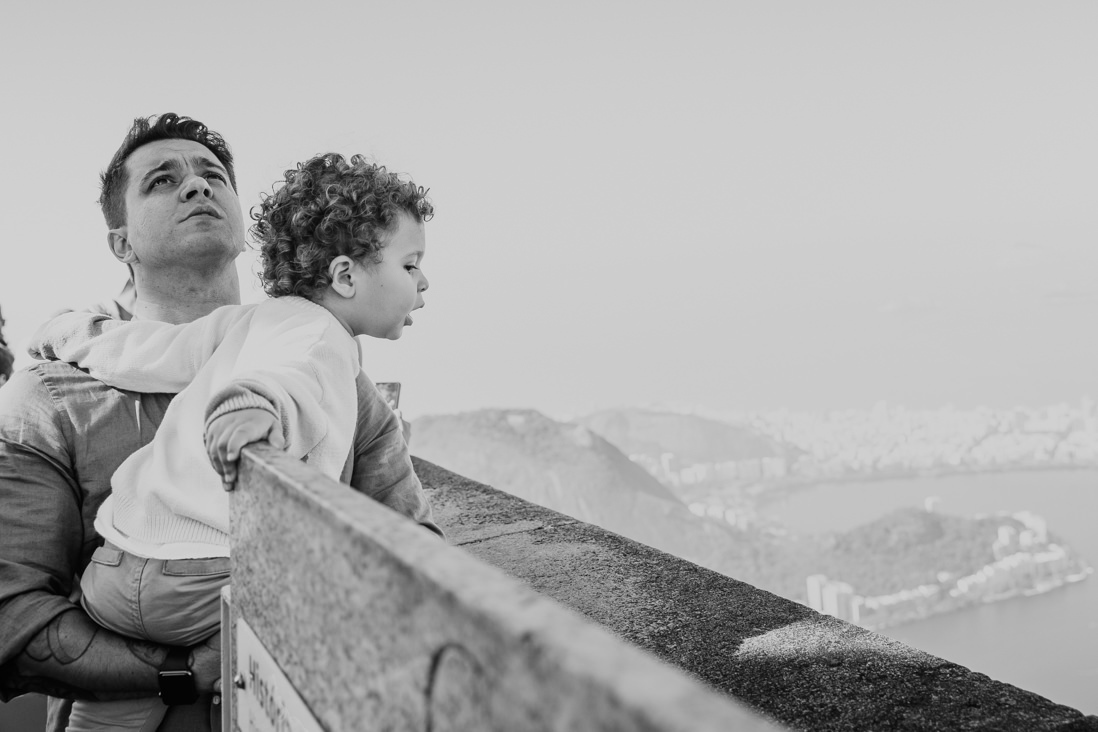 pai olha para o alto enquanto o filho olha para a baia de guanabara do alto do cristo redentor no dia do seu batizado