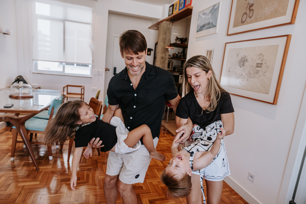 pae e mae na sala de casa brincam com os filhos pendurados na cintura de cada um