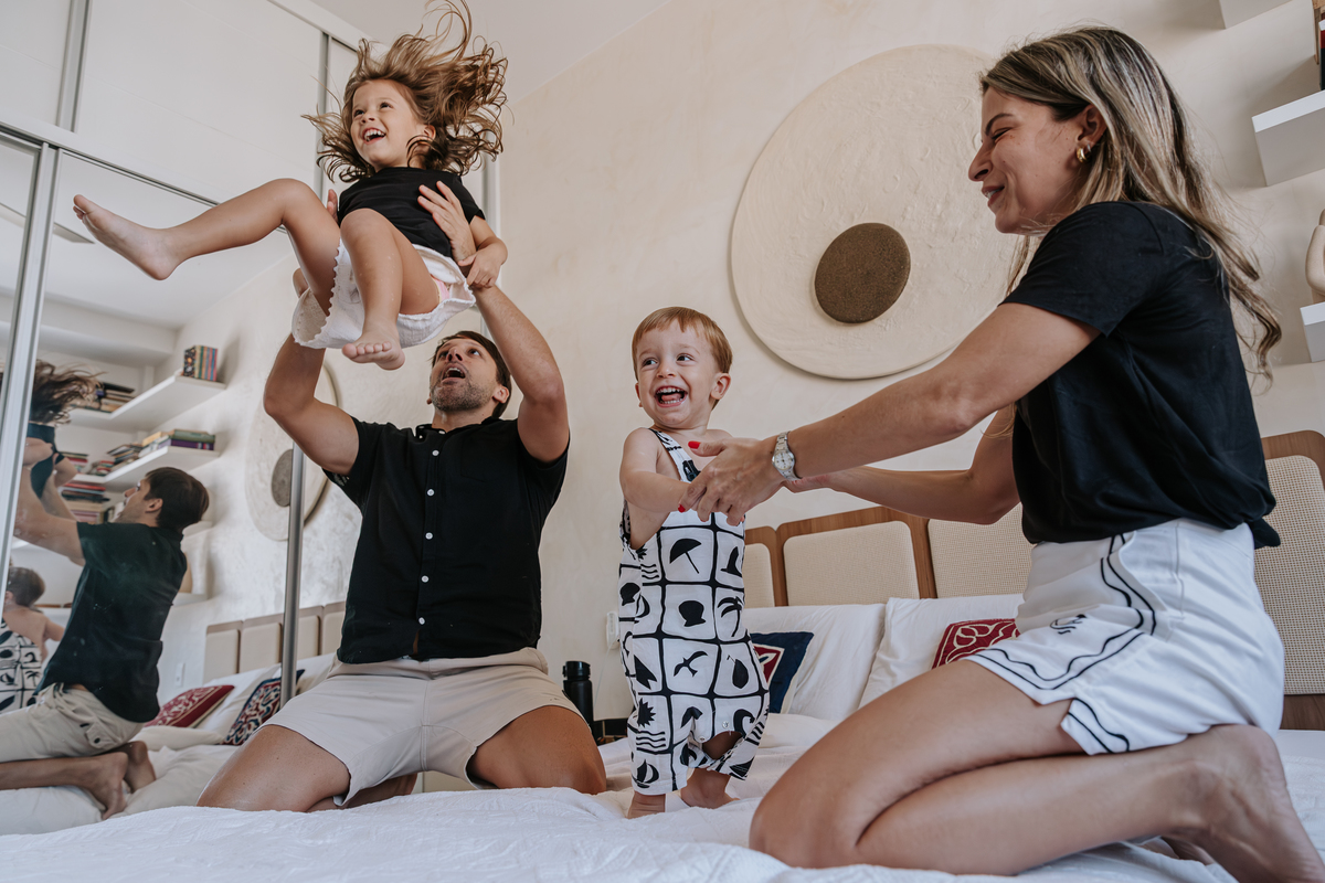 foto de uma familia de quatro pessoas fazendo bagunça na cama do casal, a filha mais velha sendo jogada para o alto por seu pai enquanto o filho mais novo de 2 anos se diverte vendo a cena