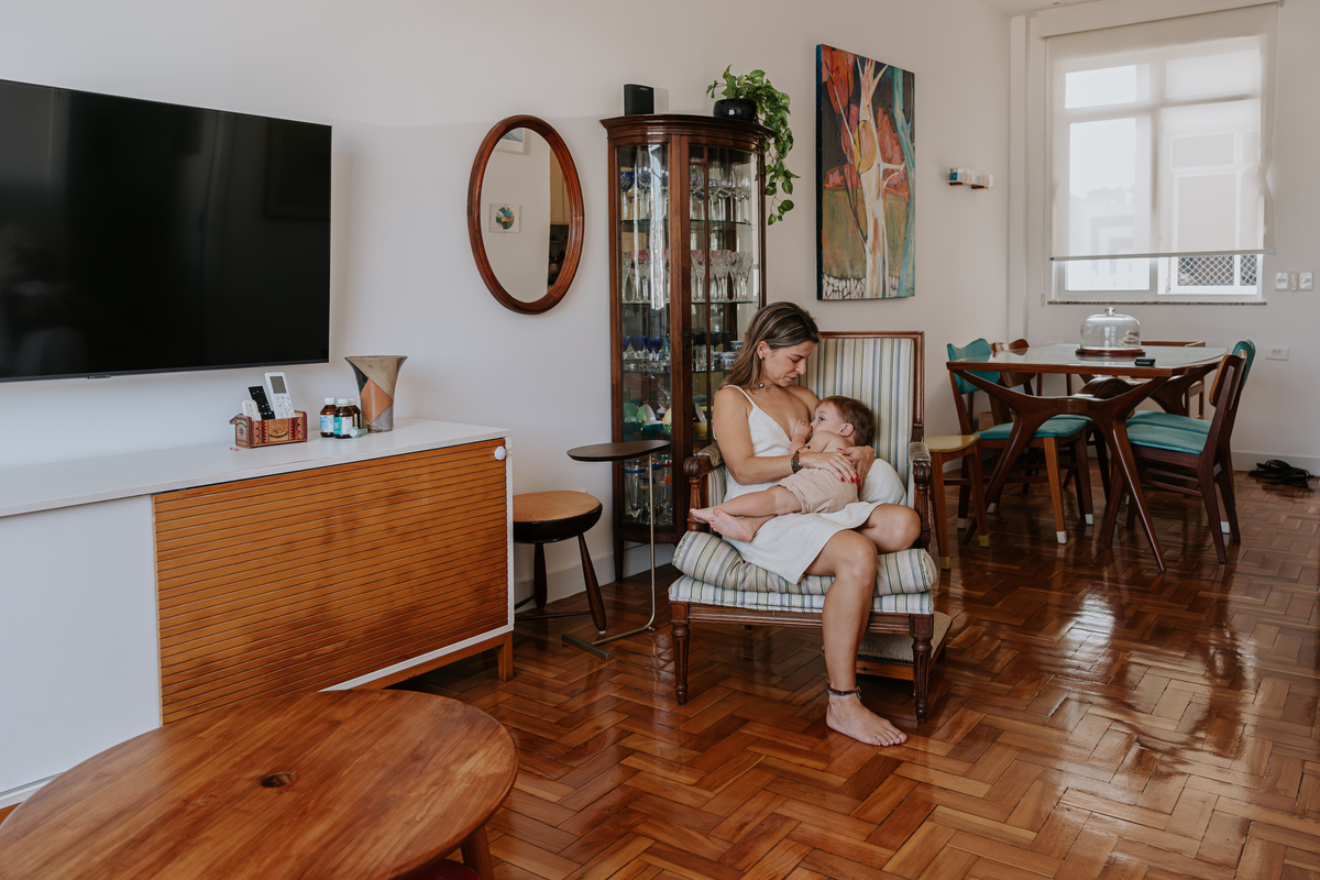 foto de plano aberto de uma sala bem iluminada com luz natural com uma mãe loira amamentando seu filho 