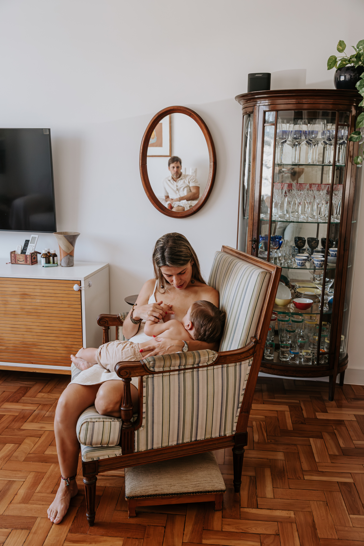 mae amamentando seu bebe sentada em uma poltrona na sala de sua casa com imagem do pai refletida num espelho