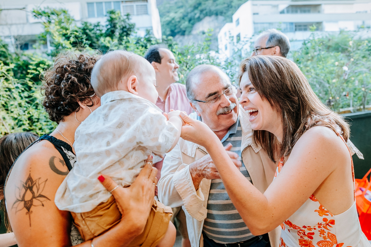Fotografia da mamae sorrindo para seu filho de um ano que esta no colo de um convidado