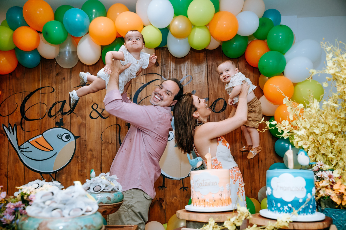 Fotografia dos pais levantando os filhos gêmeos para o alto na mesa do bolo