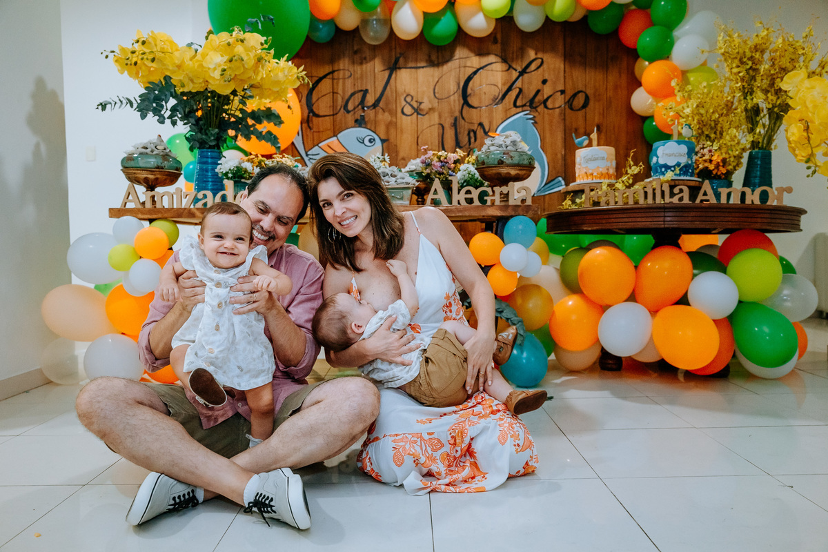 Fotografia dos pais sentados no chão com os filhos gêmeos na mesa do bolo