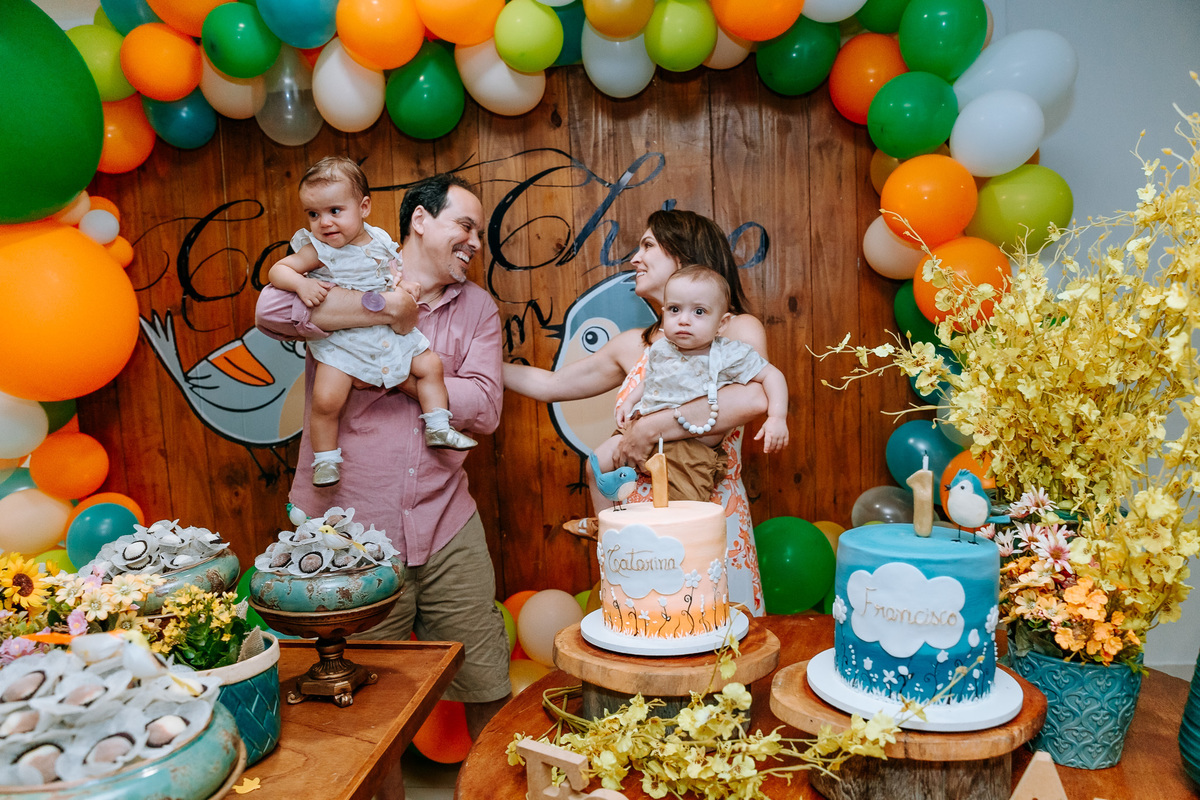 Fotografia dos pais se olhando em cumplicidade e com os gêmeos no colo na mesa do bolo na hora do parabéns