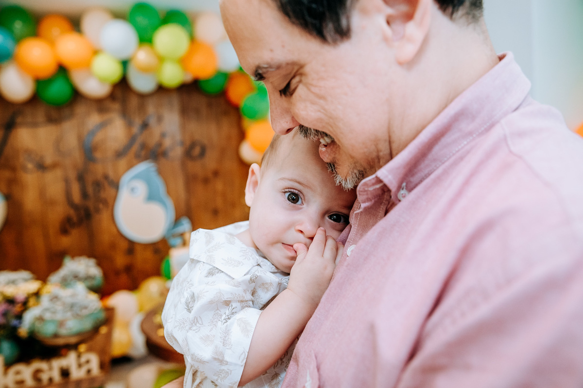 Fotografia de menino de 1 ano no colo do seu pai com a mão na boca