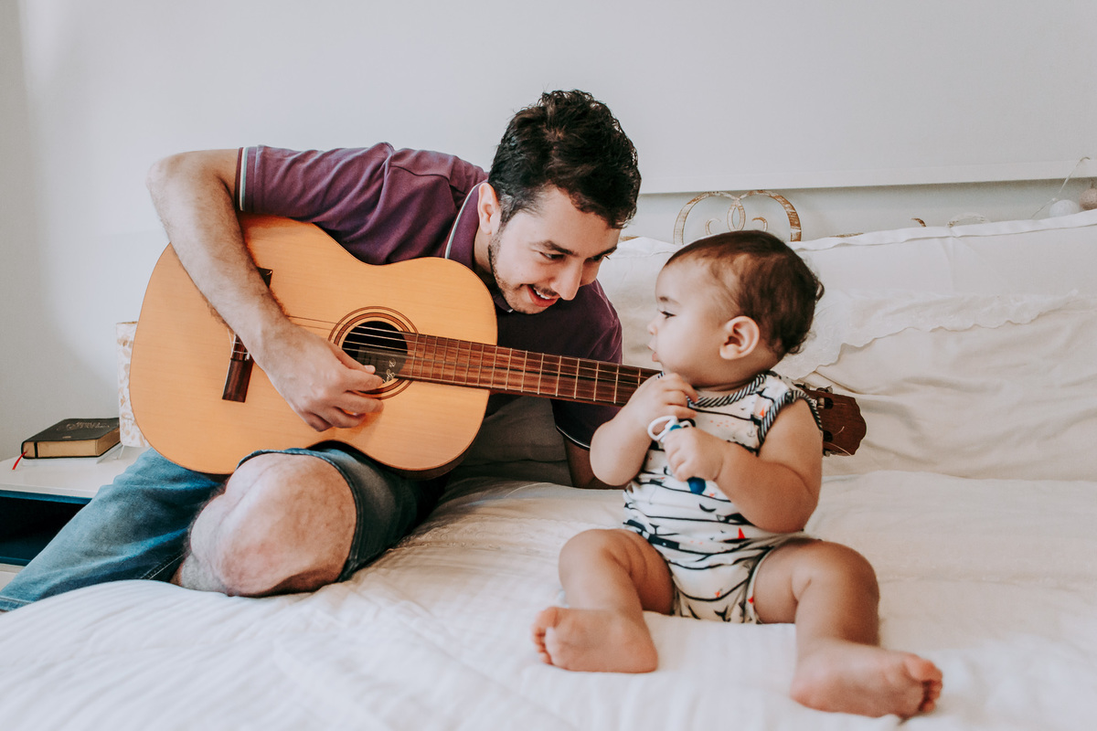 Fotografia ensaio fotográfico em casa com o pai tocando violão para o filho no quarto