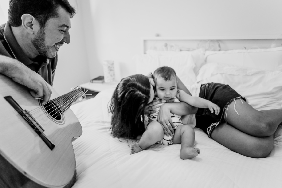 Fotografia ensaio fotográfico em casa com o pai tocando violão para mamãe e bebê