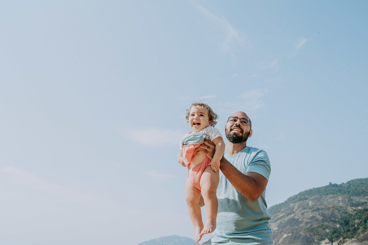 Fotografia de pai levantando a filha para o alto durante ensaio fotográfico de família na Praia Vermelha no RJ