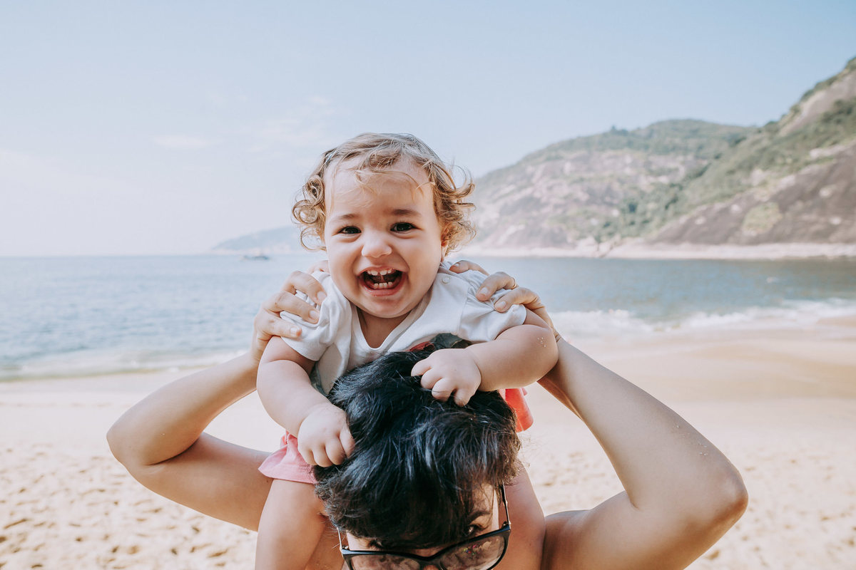 Fotografia de menina sorrindo sentada na carcunda mãe e puxando o cabelo dela olhando  para a câmera durante ensaio fotográfico na Praia Vermelha no RJ