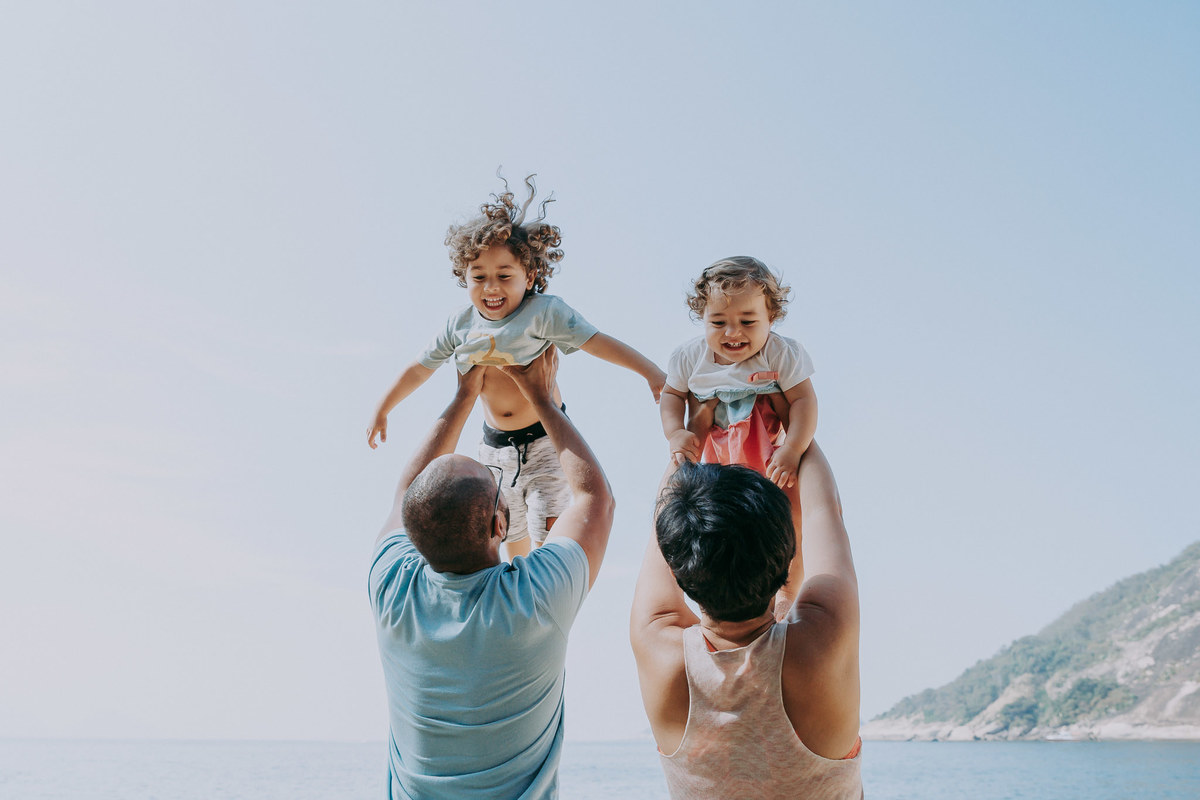 Fotografia de pai e mãe levantando os filhos para o alto durante ensaio fotográfico de família na Praia Vermelha no RJ