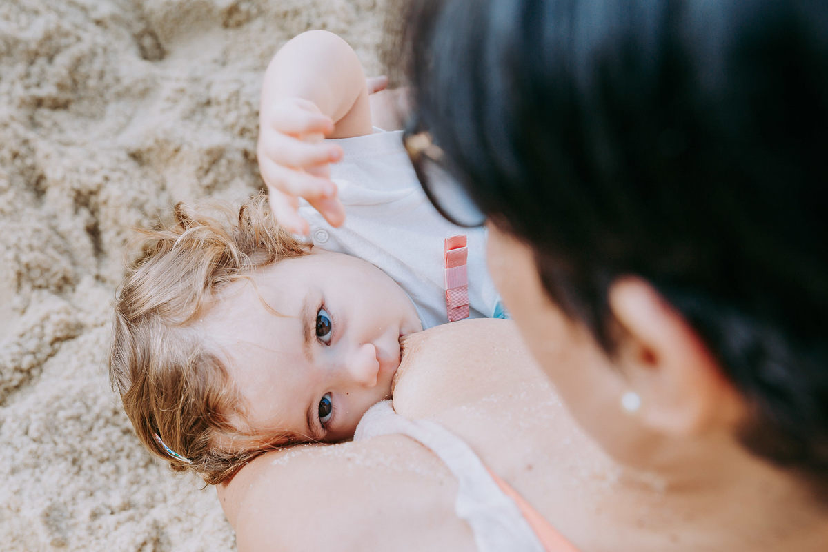 Fotografia de menina mamando na mãe durante ensaio fotográfico de família  na Praia Vermelha no RJ