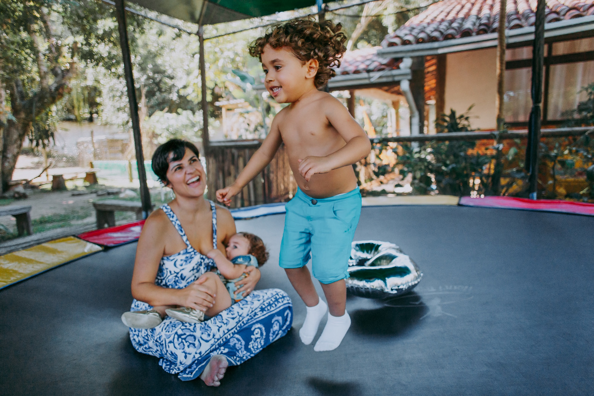 fotografia de mae olhando admirada para o filho no pula pula enquanto amamenta o outro