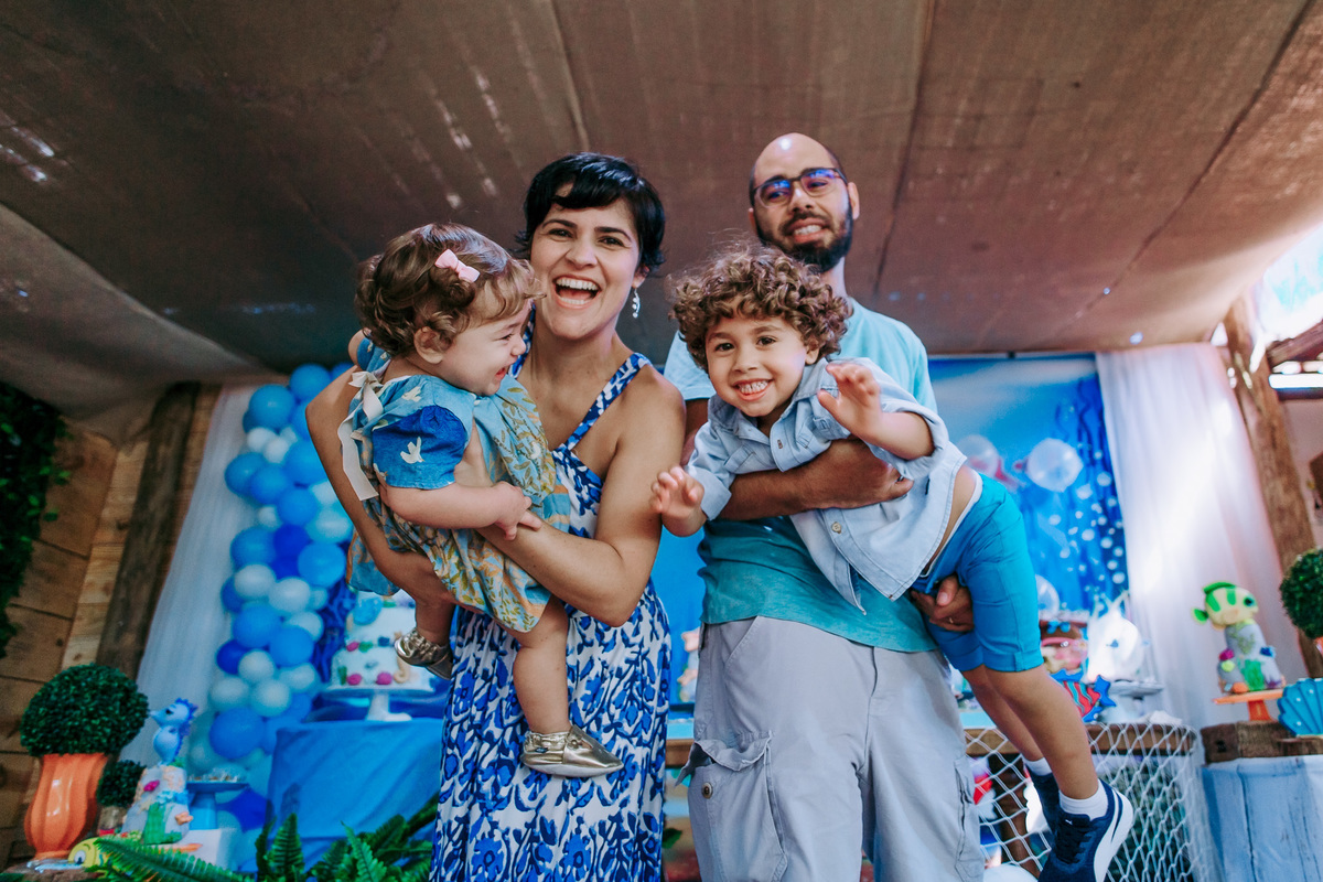 fotografia dos pais fazendo aviaozinho com os dois filhos em frente a mesa do bolo