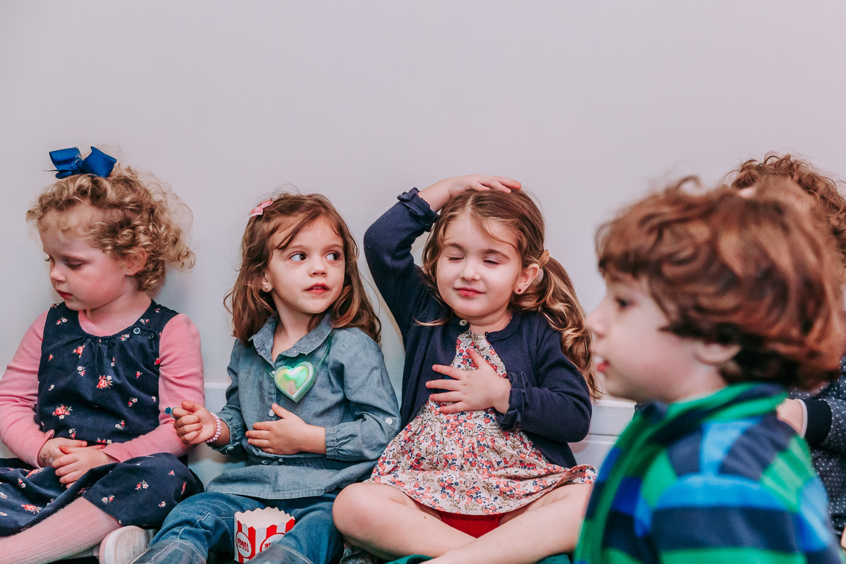 fotografia de crianças sentadas no chao durante recreaçao infantil em festa de aniversario, uma delas está de olhos fechados com a mao na cabeça e outra no peito