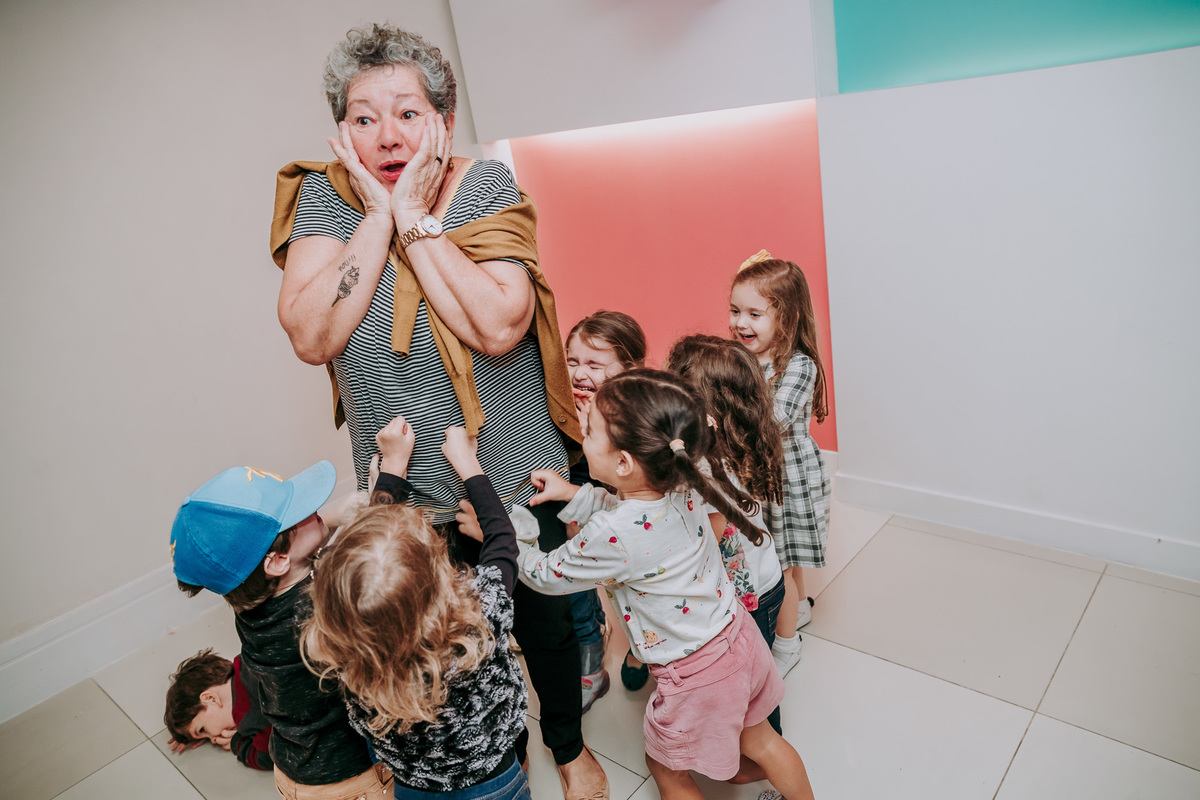 fotografia de geracoes em que crianças brincam puxando a roupa de senhora que está com mao no rosto fazendo cara de assustada 