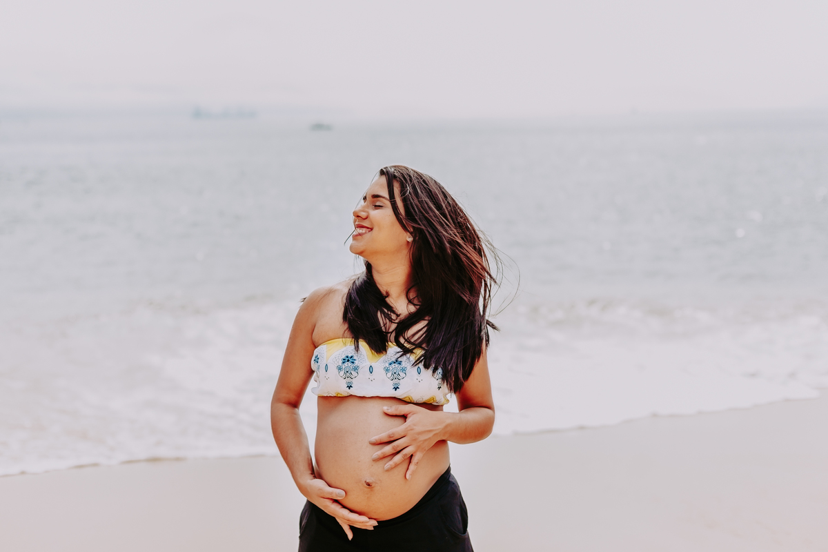 gestante sacode o cabelo em frente ao mar sorrindo e segurando a sua barriga