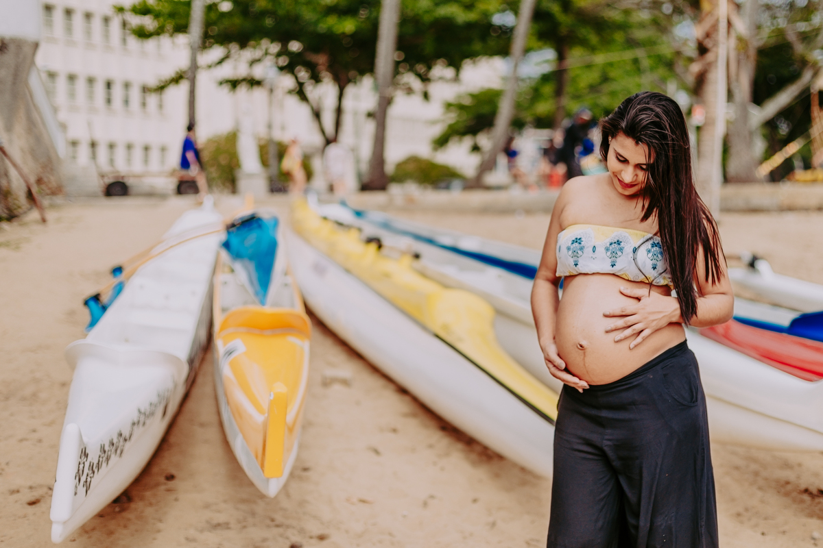 gestante posando com a mao na barriga e olhando para barriga em frente a varios caiaques na praia vermelha