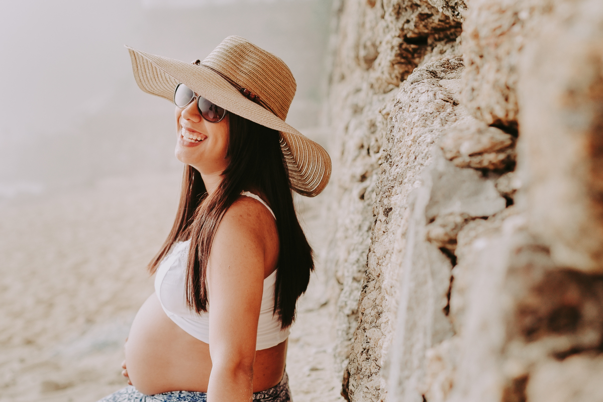 gestante sentada em pedras de chapeu e oculos escuros sorrindo