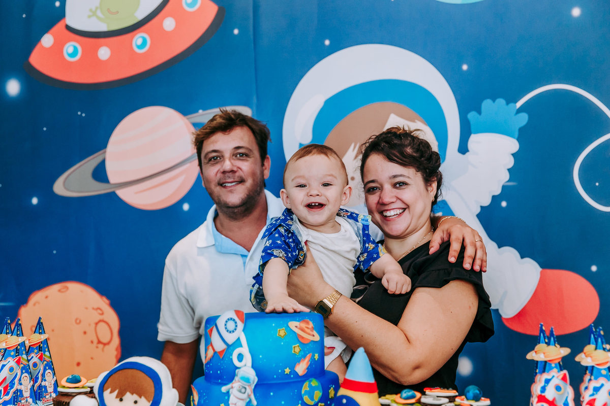 foto do aniversariante sorrindo posando com padrinhso na mesa do bolo e batendo no bolo