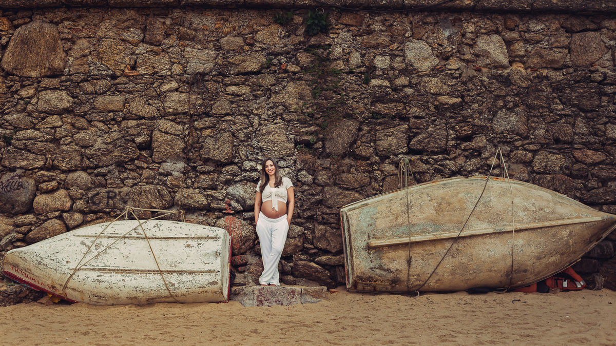 foto de gestante de corpo inteiro encostada num paredão de pedras entre dois barcos na praia vermelha no rio de janeiro