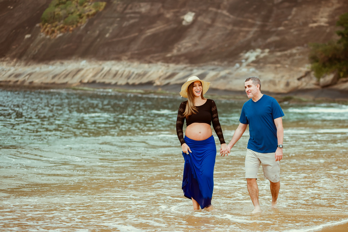 Foto de gestante e marido caminhando de mãos dadas pela praia vermelha